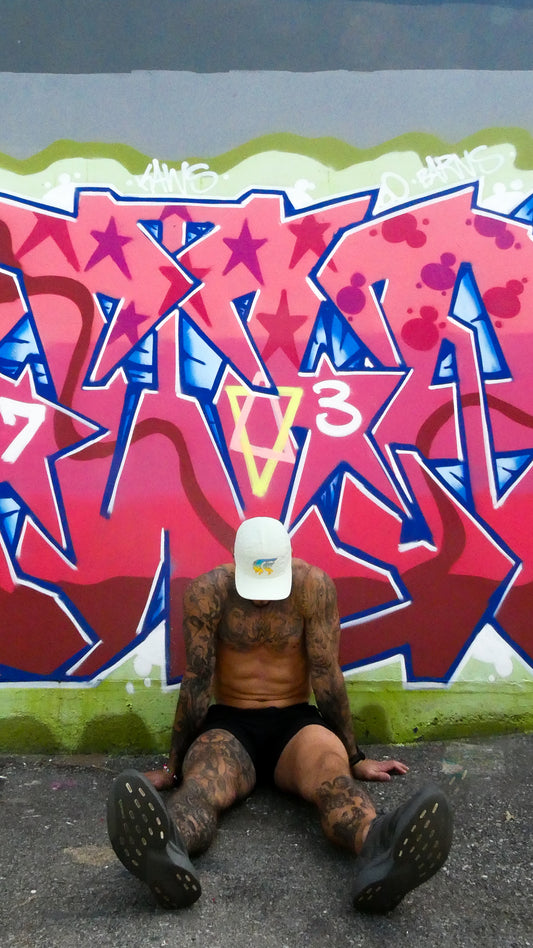 Person sitting on the ground with a white cap in front of a colorful graffiti wall.